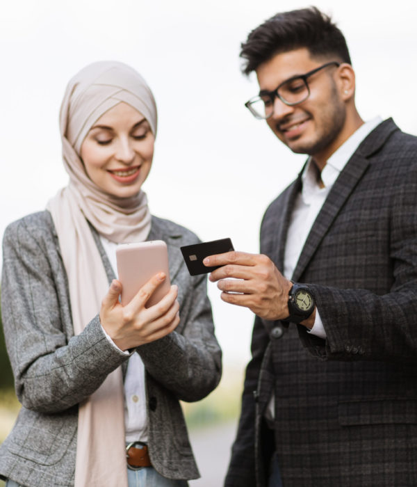 Conceptual photo of hindu businessman showing credit card number to his female Muslim colleague typing on smartphone and making online payment. Happy people outdoors shopping online.